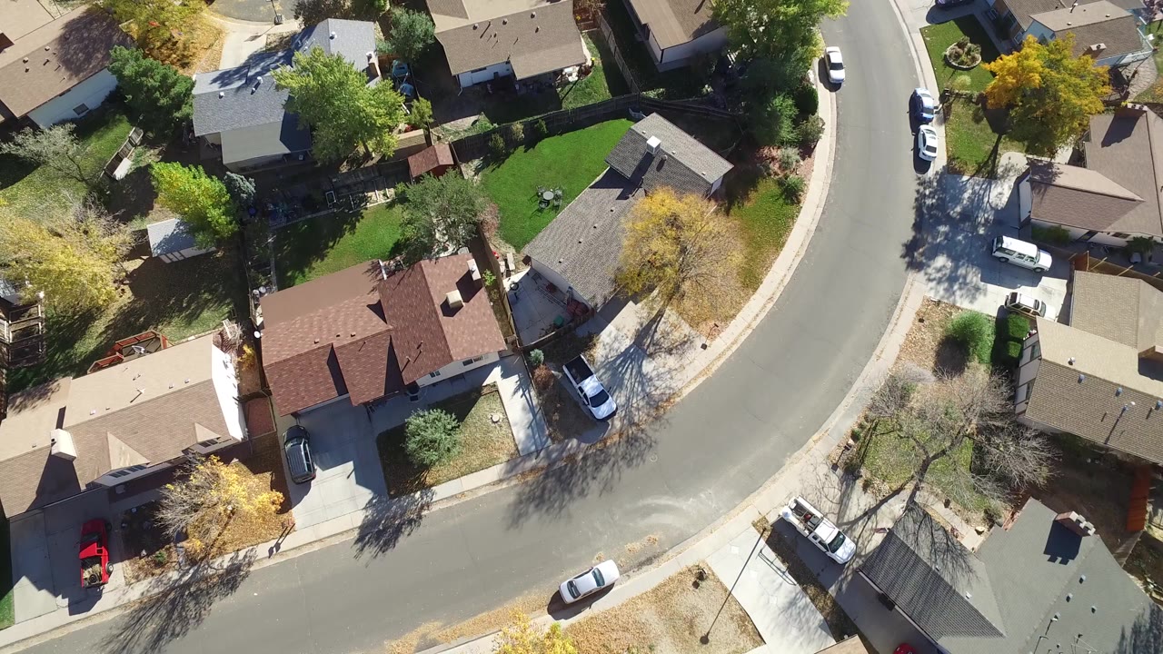 Overhead drone frame in Hidden Lake, Colorado showing a curved residential street bordered by single-family homes, fenced yards, green lawns, parked cars, and no visible people.