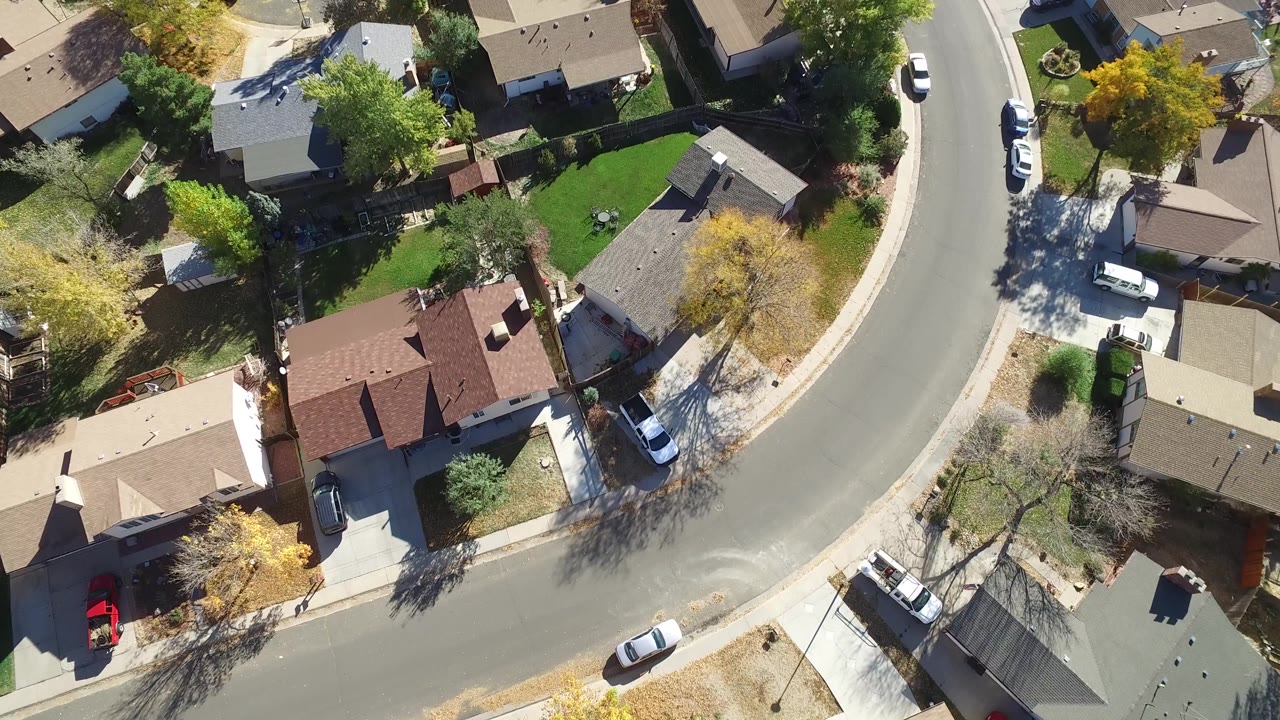 Hidden Lake, Colorado aerial frame with the road curve shifted through the center, showing detached homes, driveways, lawns, autumn trees, parked vehicles, and no visible people.