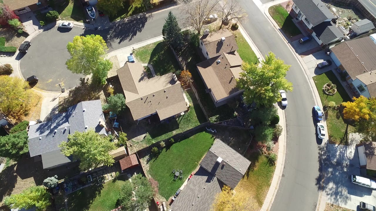 Opening overhead frame of Hidden Lake, Colorado homes beside curved residential streets, with green lawns, mature trees, parked cars, strong sun, and no visible people.