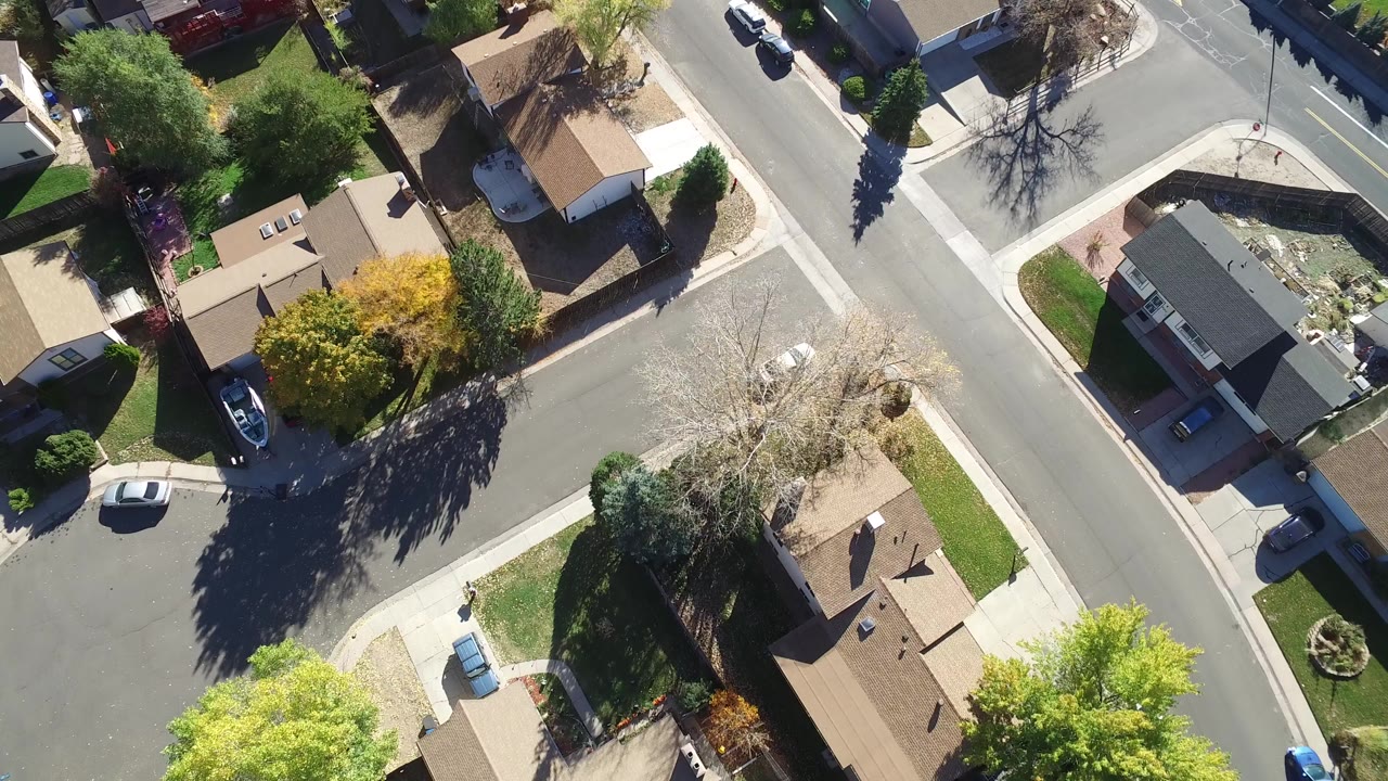 Opening overhead frame of Hidden Lake, Colorado showing a residential street corner, detached homes, green yards, trees, driveways, parked cars, and no visible people.