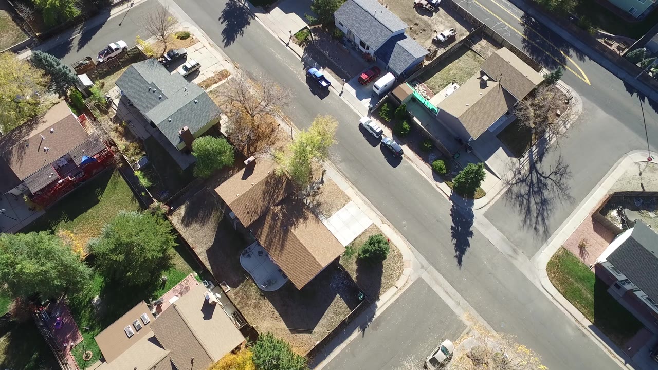 Second overhead frame of Hidden Lake, Colorado showing rooftops, a residential road, driveways, parked cars, small yards, and strong tree shadows across pavement.