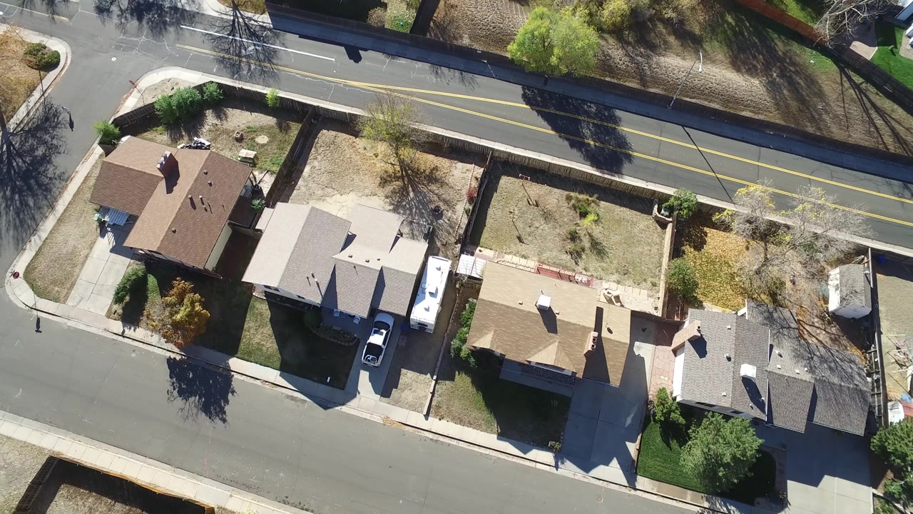 Overhead frame in Hidden Lake, Colorado with rooftops filling the lower half, a parked white vehicle in a driveway, dry yards, sidewalks, and the main road running horizontally near the top.