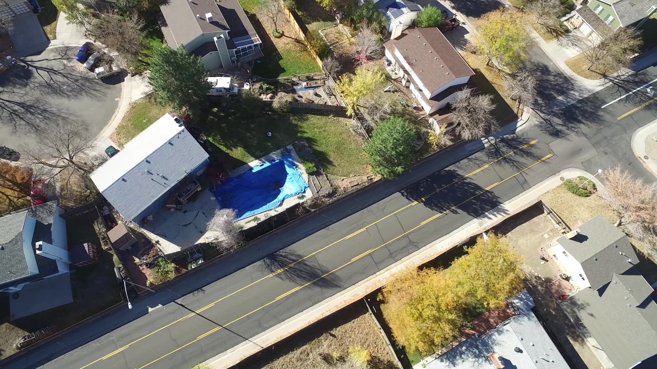Aerial frame in Hidden Lake, Colorado with a road through the center, flat and pitched rooftops, tree shadows, a blue covered yard feature, and no visible people.