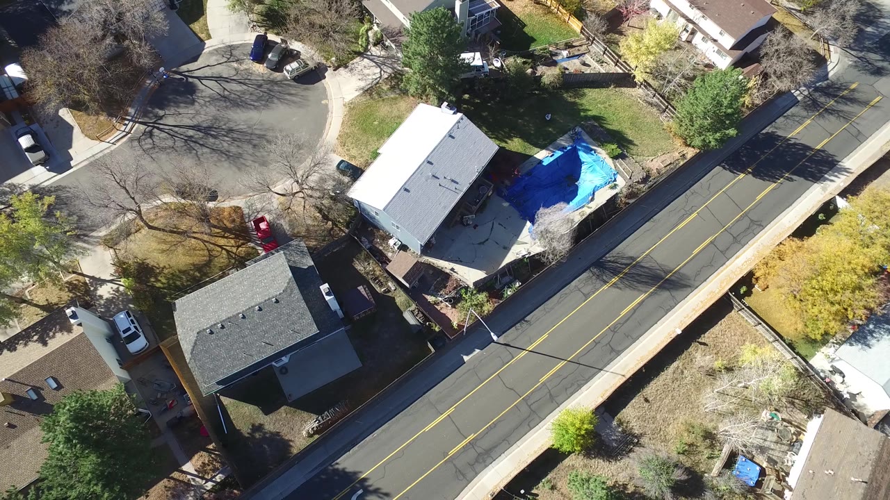 Aerial frame over Hidden Lake, Colorado with a diagonal residential road, a white-roofed building, adjacent homes, yards, trees, and no visible people.
