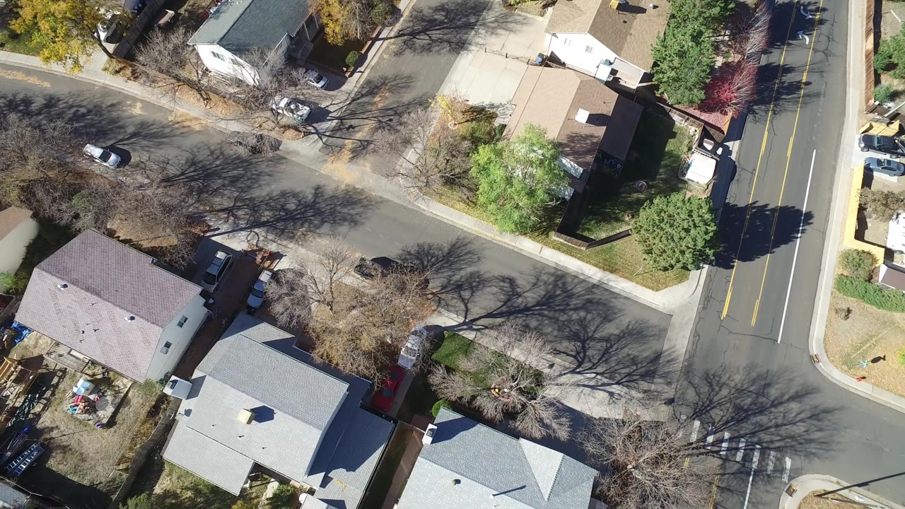 Aerial frame in Hidden Lake, Colorado with gray and brown rooftops, a curved side street, a wider marked road at right, parked cars, yard fencing, and no clearly visible people.