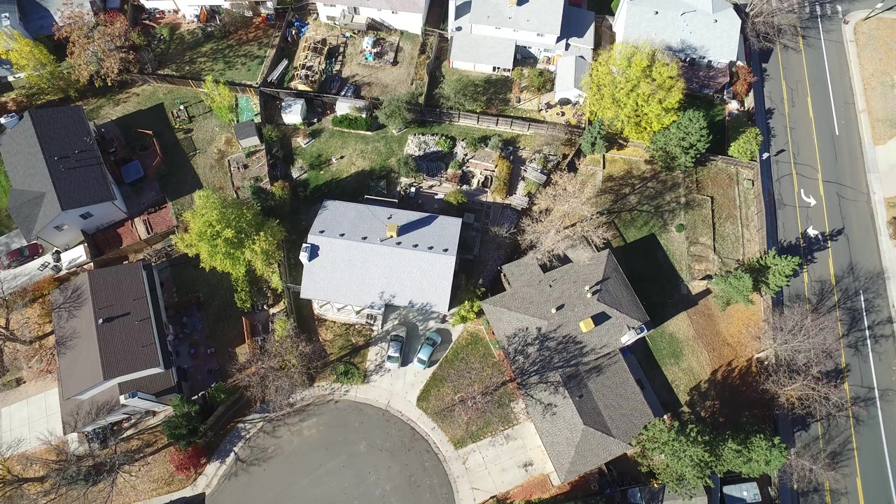Hidden Lake, Colorado aerial frame with the same residential block centered on pale and dark roofs, small yards, driveways, parked vehicles, and the road at frame right.