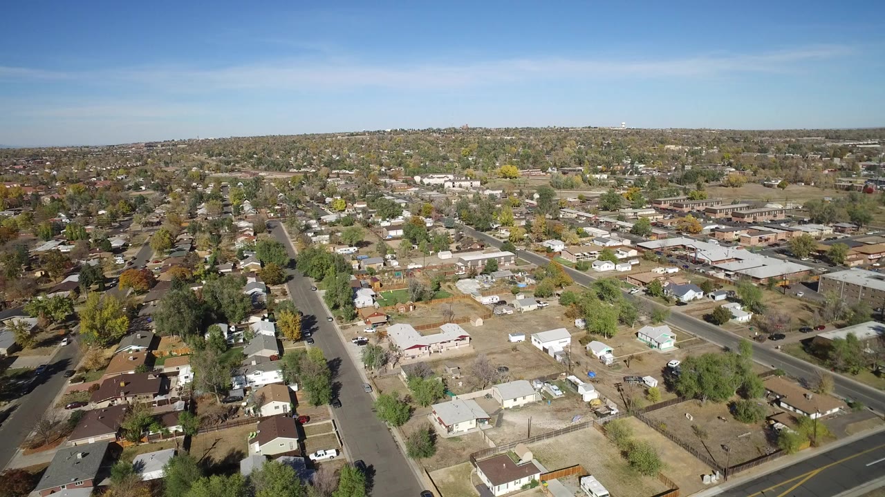 High aerial view over houses, streets, trees, and dry yards under blue sky.