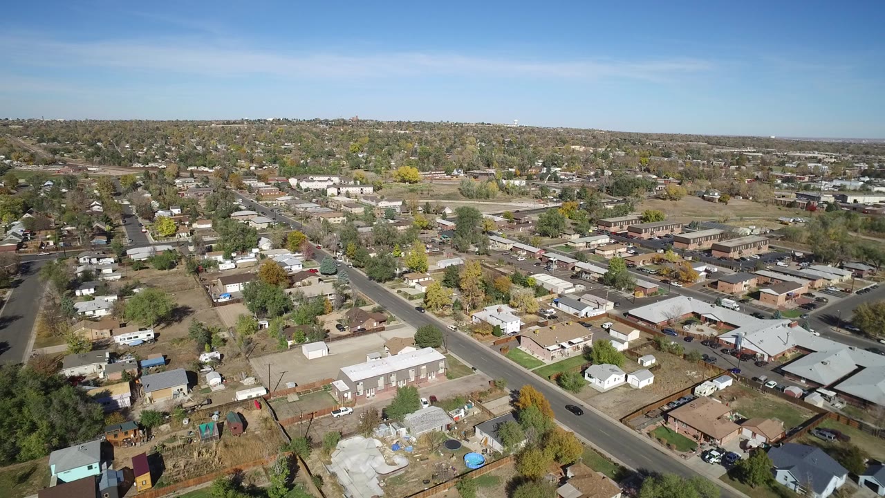 Opening aerial frame over Hidden Lake, Colorado, with single-story homes, a diagonal road, trees, distant low buildings, and no visible people.
