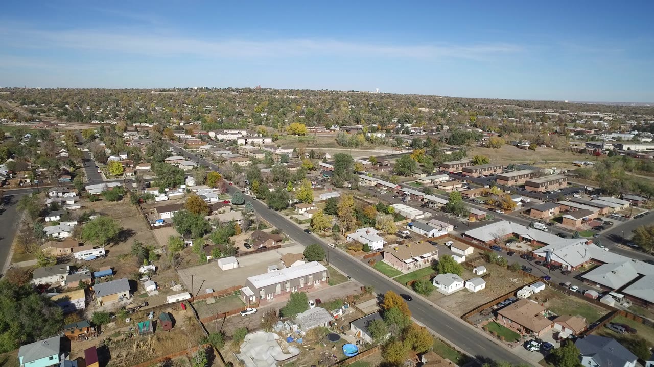 Suburban aerial with a diagonal street, rooftops, yards, and distant development.