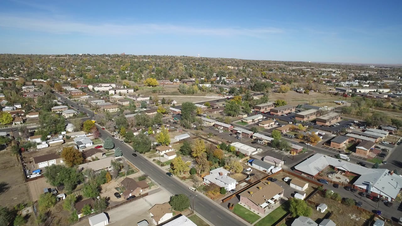 Opening aerial frame over Hidden Lake Colorado with a diagonal road, low homes, scattered trees, distant hills, clear blue sky, and no visible people.