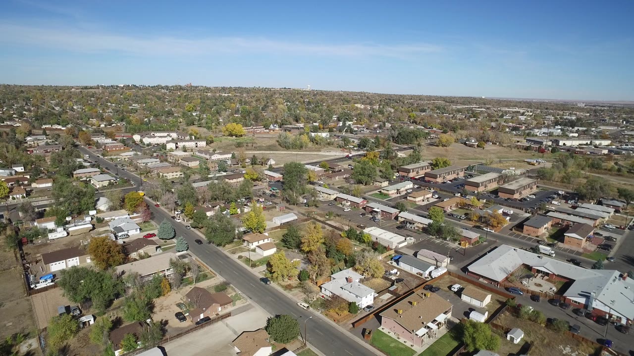 Aerial neighborhood view with streets, tan and gray rooftops, trees, and hills on the horizon.