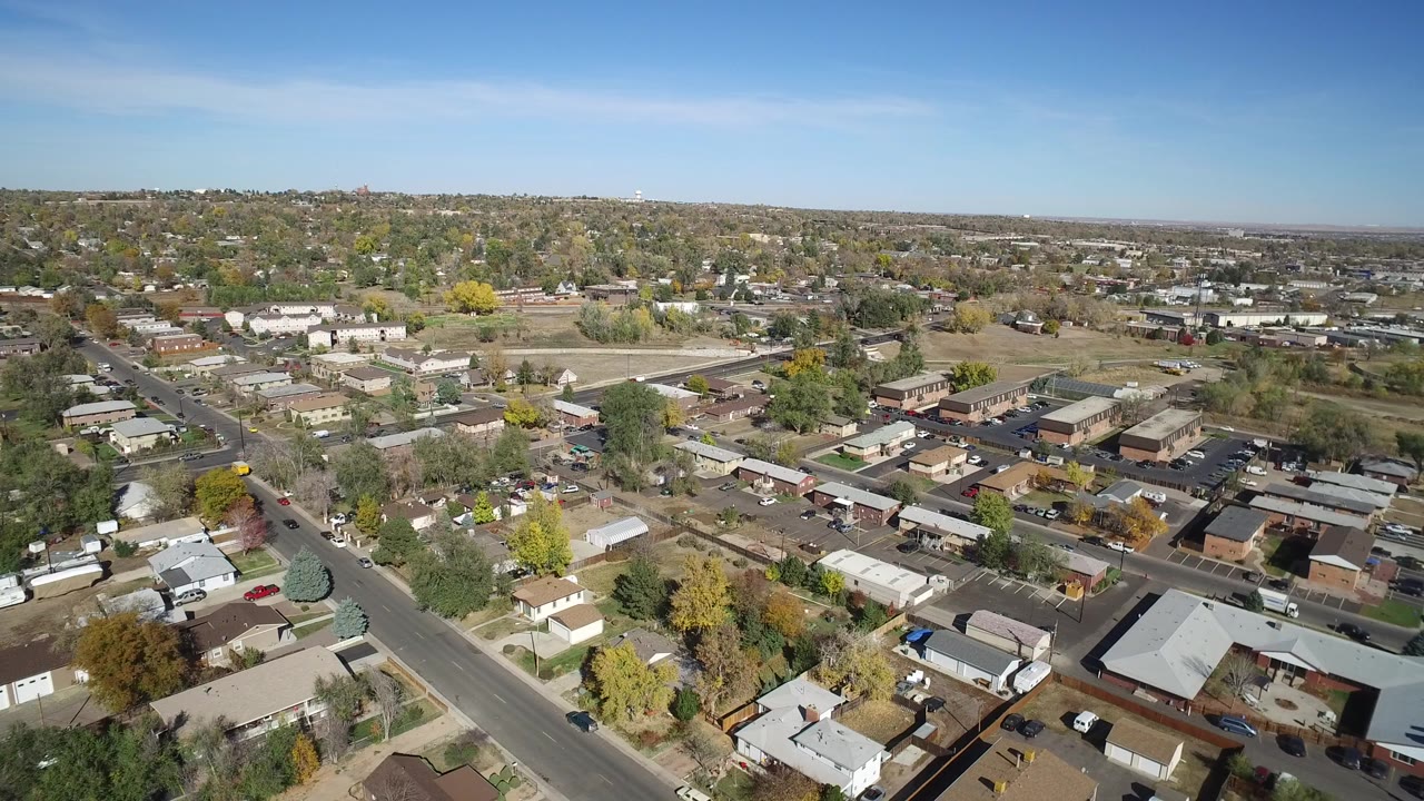 High aerial view over roads, houses, low buildings, trees, and blue sky.