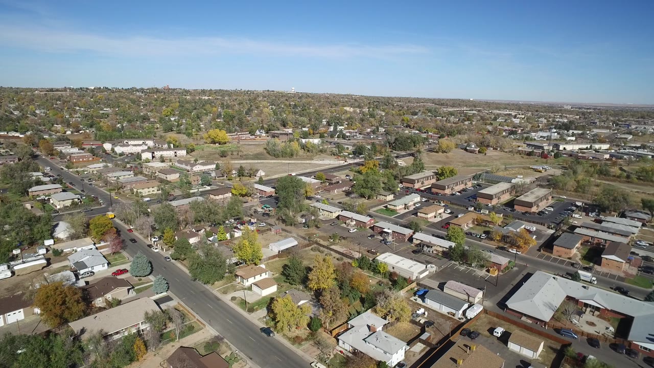 High oblique aerial frame over Hidden Lake, Colorado, with a diagonal road at left, small buildings in the center, tree-lined lots, distant development, and no visible people.
