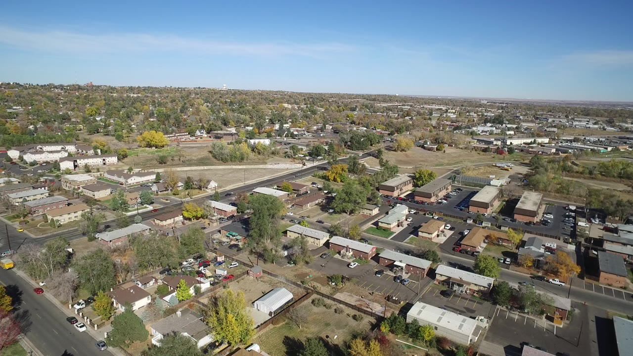 Aerial view over homes, roads, small buildings, trees, and open lots under a blue sky.