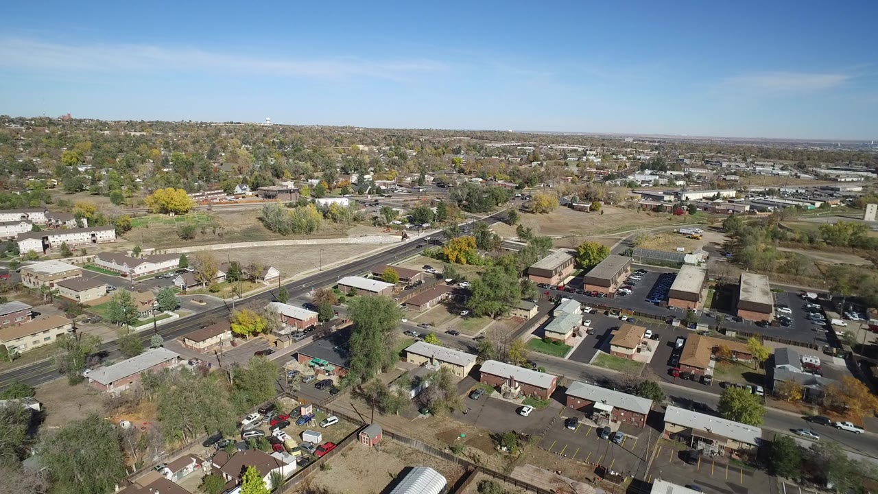 High aerial view over Hidden Lake, Colorado, with a diagonal main road, low homes, parked cars, dry lots, scattered trees, and no visible people.