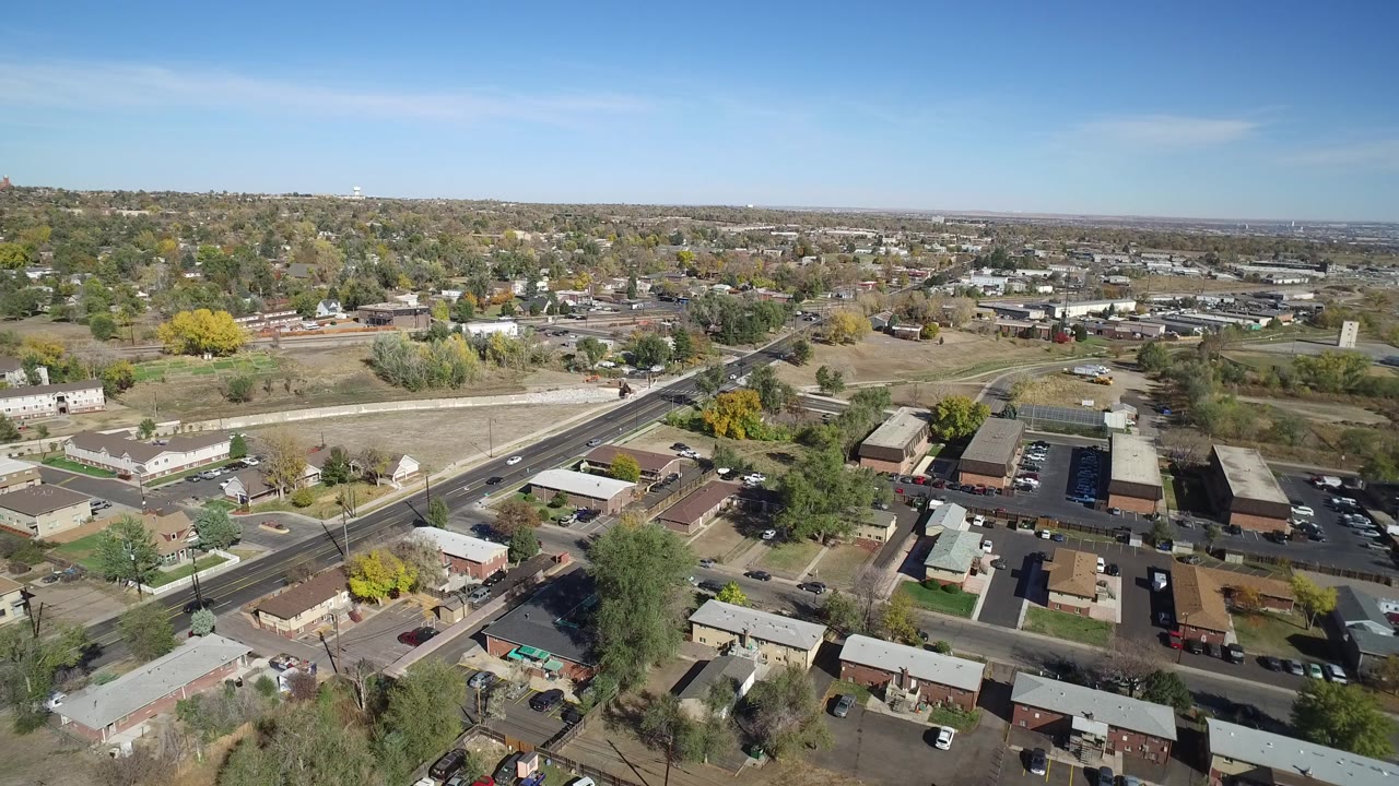 Early aerial frame over Hidden Lake, Colorado, with a diagonal road, nearby rooftops, open ground, distant town blocks, clear blue sky, and no visible people.