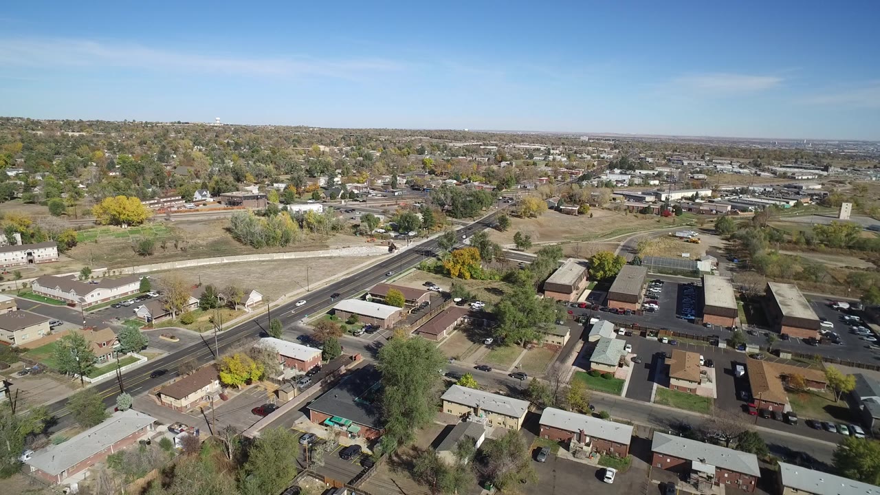 Mid-clip aerial view of Hidden Lake, Colorado, with a sunlit road, residential roofs, industrial-style buildings, open lots, distant neighborhoods, and no visible people.
