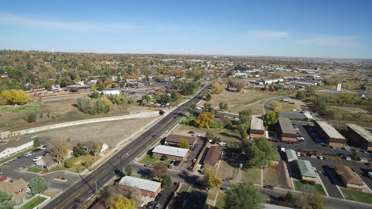 Aerial view over a road, houses, trees, and open lots under blue sky.
