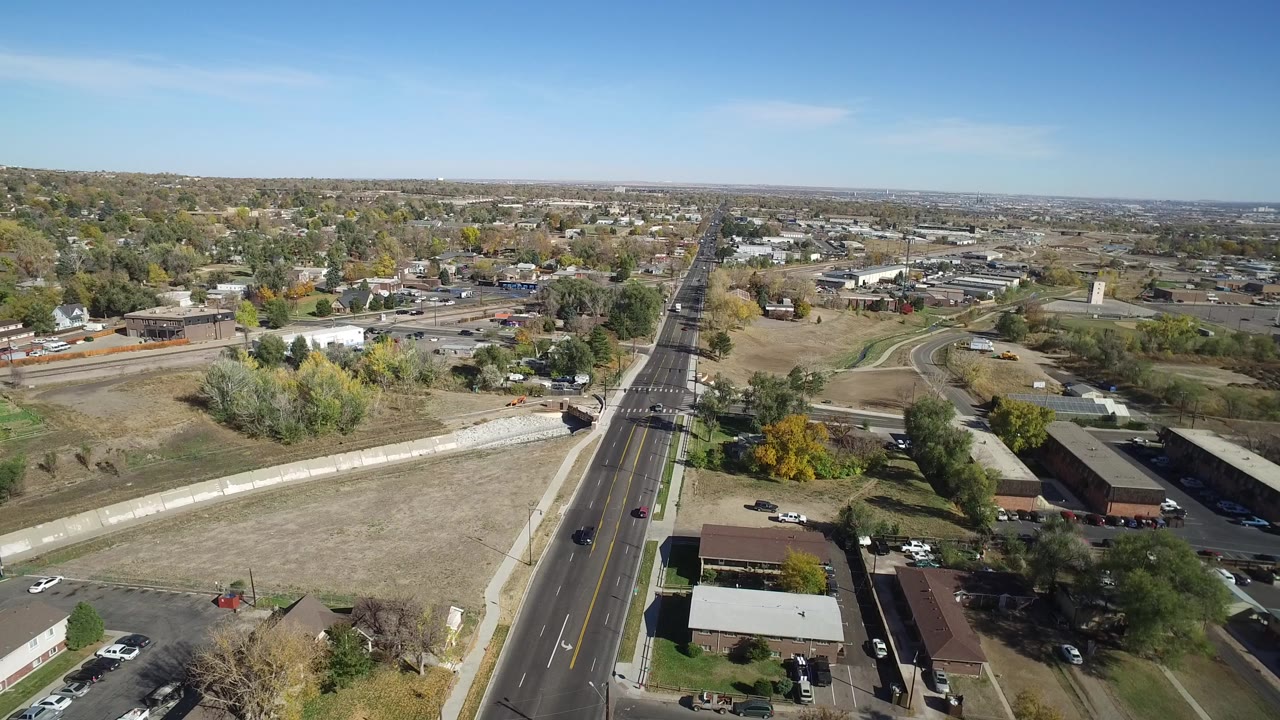 Opening aerial frame over Hidden Lake, Colorado, showing a straight multi-lane road, dry fields, low buildings, scattered trees, distant neighborhoods, and no visible people.
