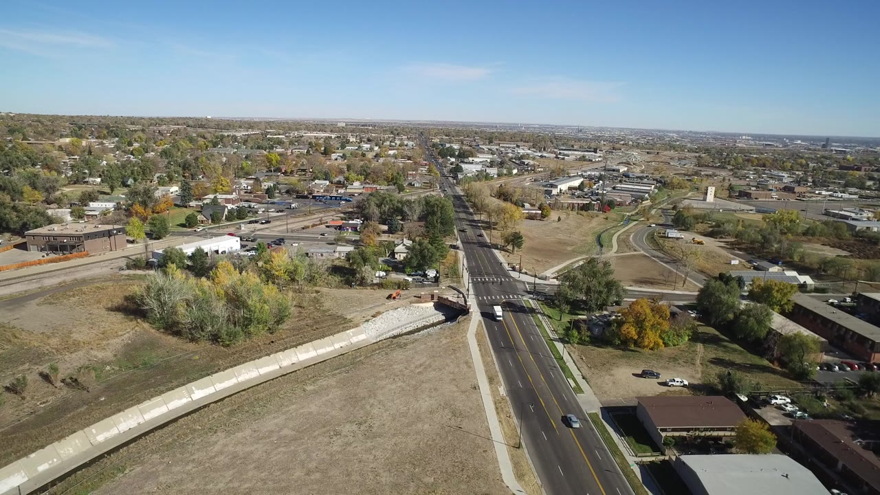 Aerial view over a road, drainage channel, trees, homes, and low buildings.