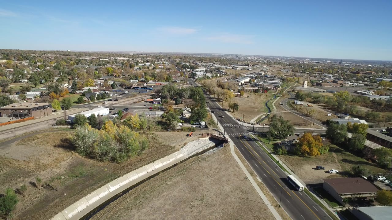 Aerial view of a road crossing, canal, open fields, homes, and distant suburbs under blue sky.