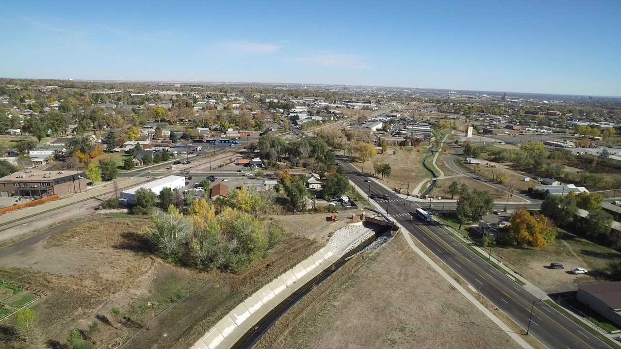Aerial view of roads, homes, trees, open lots, and drainage channels under a clear sky.