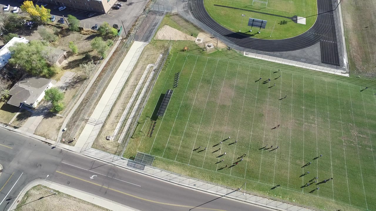 Top-down field view with sidewalks, road, and track at the edges.