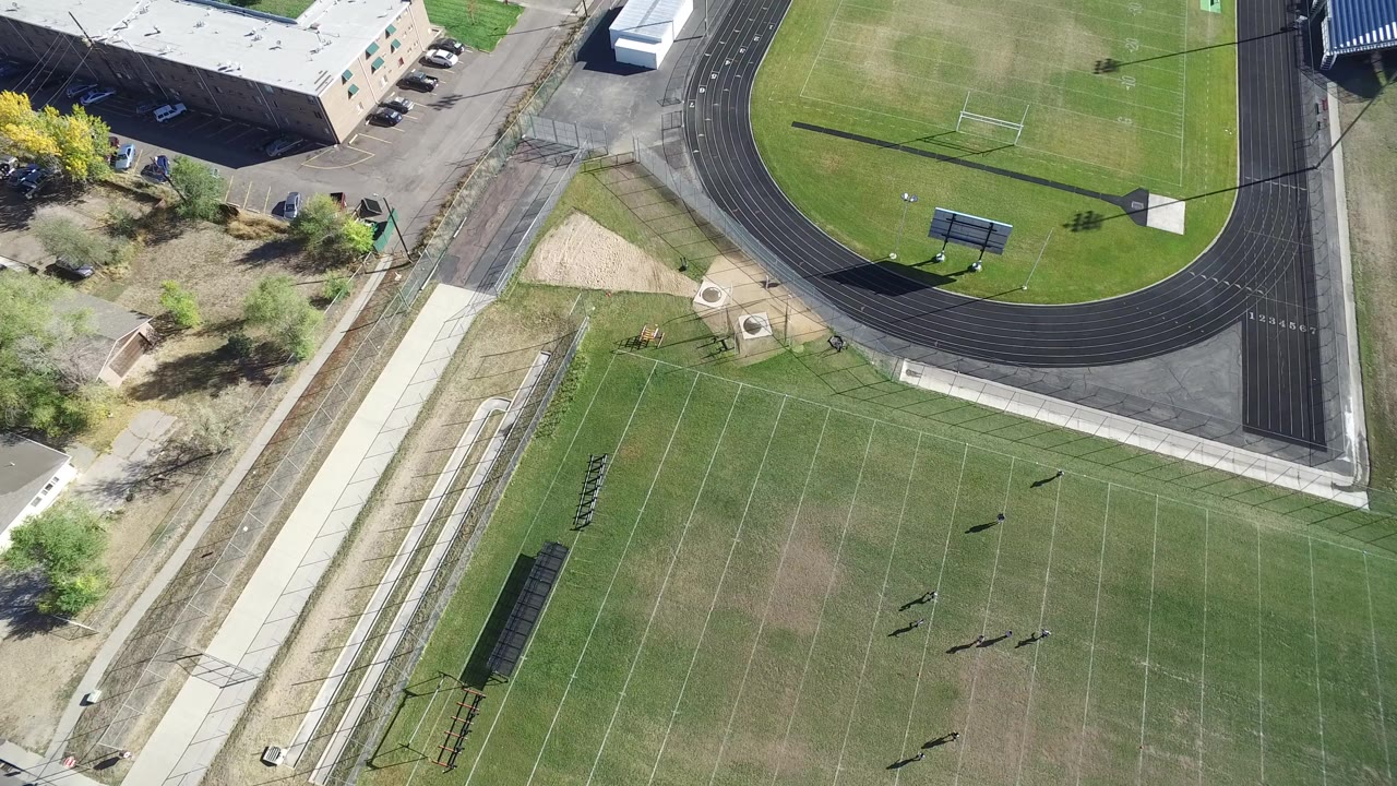 High aerial frame over a Hidden Lake, Colorado school athletic complex, with a black running track, marked football field, campus building, sidewalks, and no visible people.