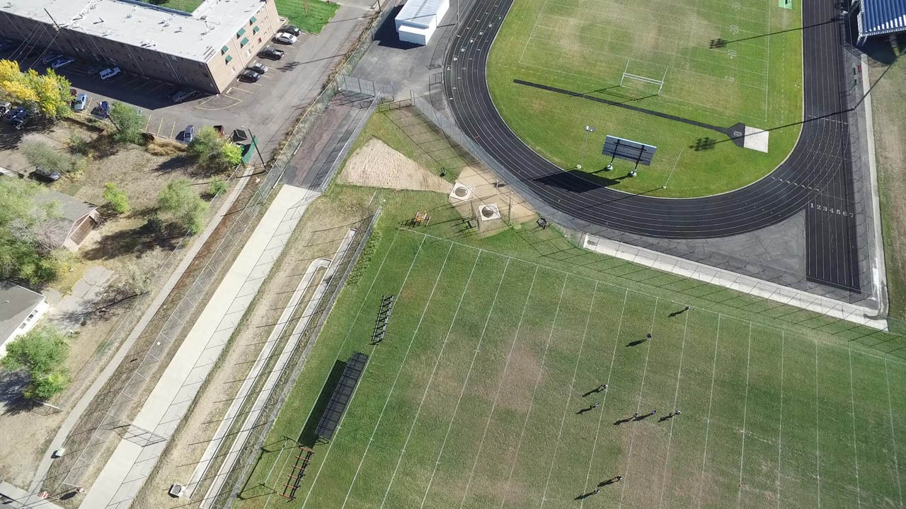 Overhead Hidden Lake, Colorado view with the track curve at upper right, the football field below, bleachers near the sideline, dry ground patches, and no visible people.