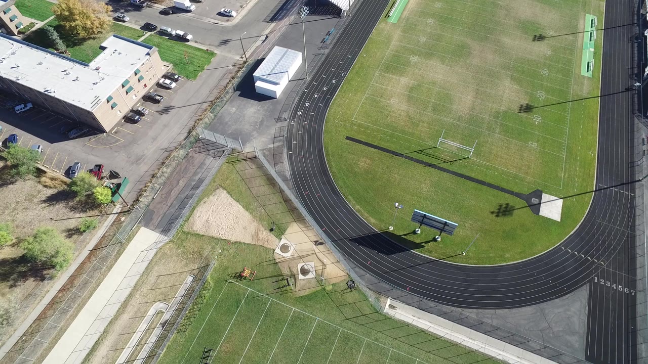 High aerial view in Hidden Lake, Colorado showing the lower curve of a black running track around a green football field, with nearby buildings, parking lots, sidewalks, and no visible people.
