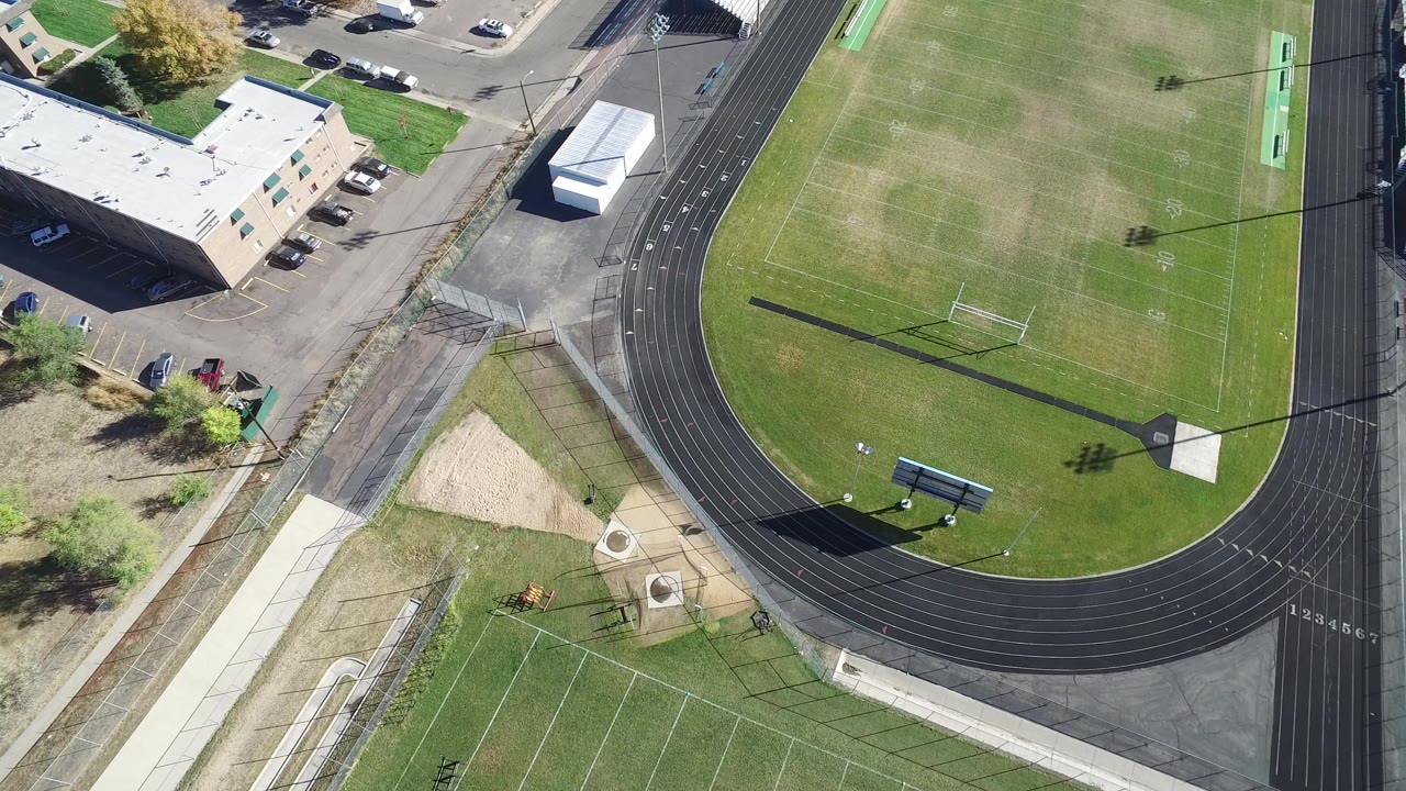 Overhead frame from Hidden Lake, Colorado with the football field shifted slightly across the view, showing the track curve, goal post, light pole shadow, adjacent pavement, and no visible people.