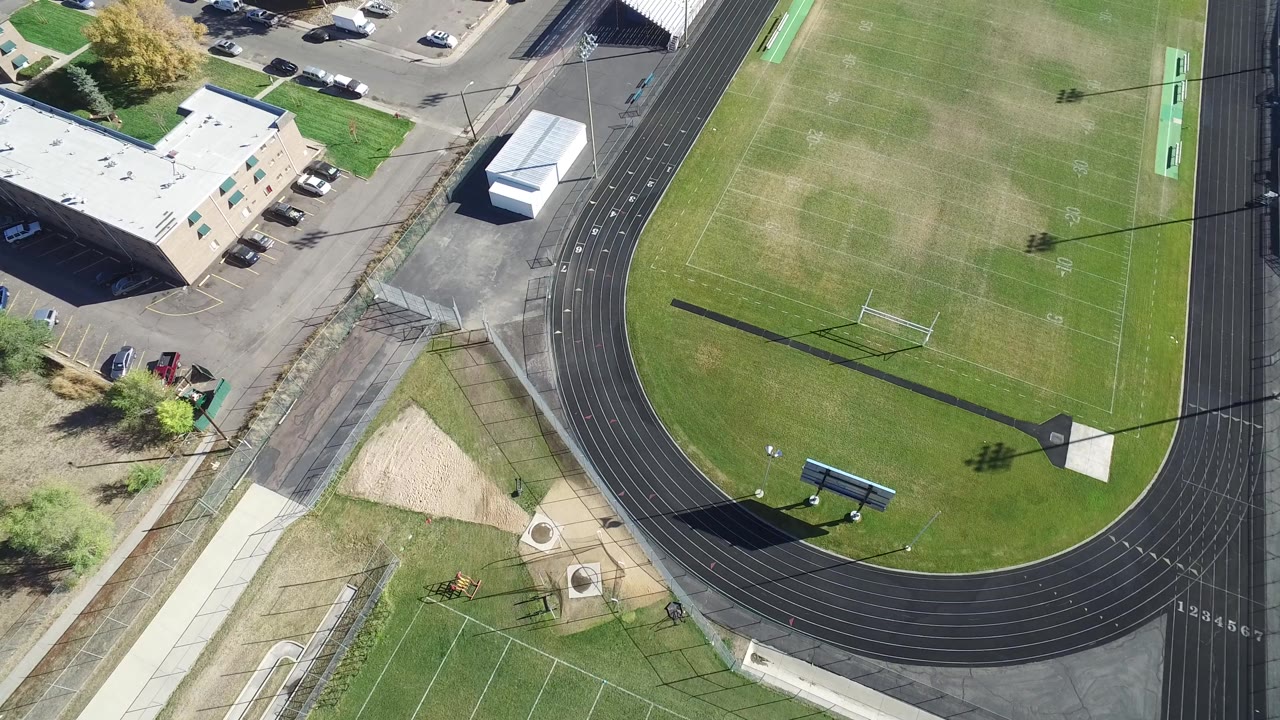 Aerial frame in Hidden Lake, Colorado showing more of the right-side football field and black track lanes, with parked cars near buildings at the top edge and no visible people.