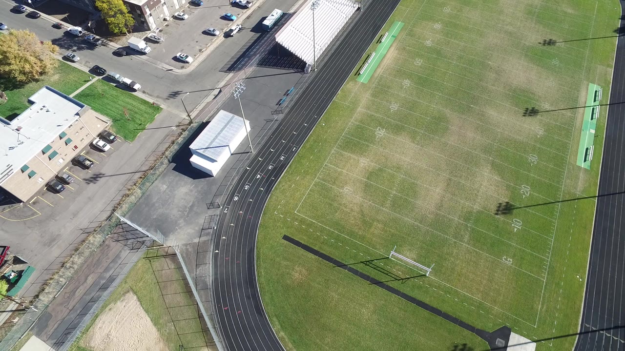 Aerial view in Hidden Lake, Colorado showing the left edge of a football field, a black running track, bleachers, nearby buildings, roads, parked cars, and no visible people.