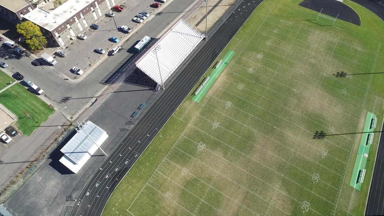 Overhead football field with track, bleachers, parked cars, and nearby buildings.