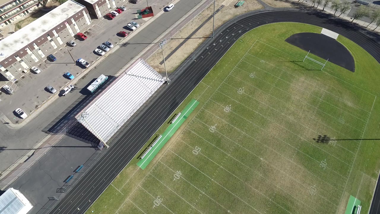 Aerial view of a football field, running track, bleachers, street, and parked cars.