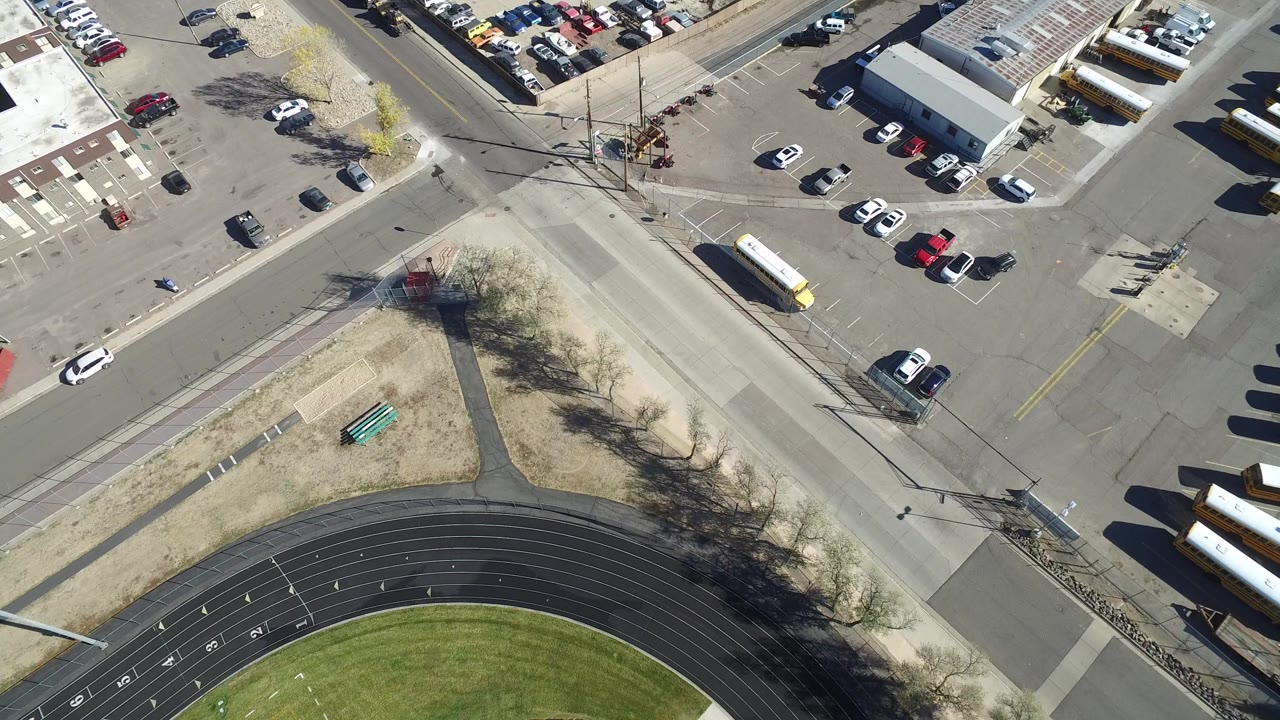 Overhead frame from Hidden Lake, Colorado showing a road intersection, curved running track, dry grass, parked cars, buses, and no visible people at the start of the clip.