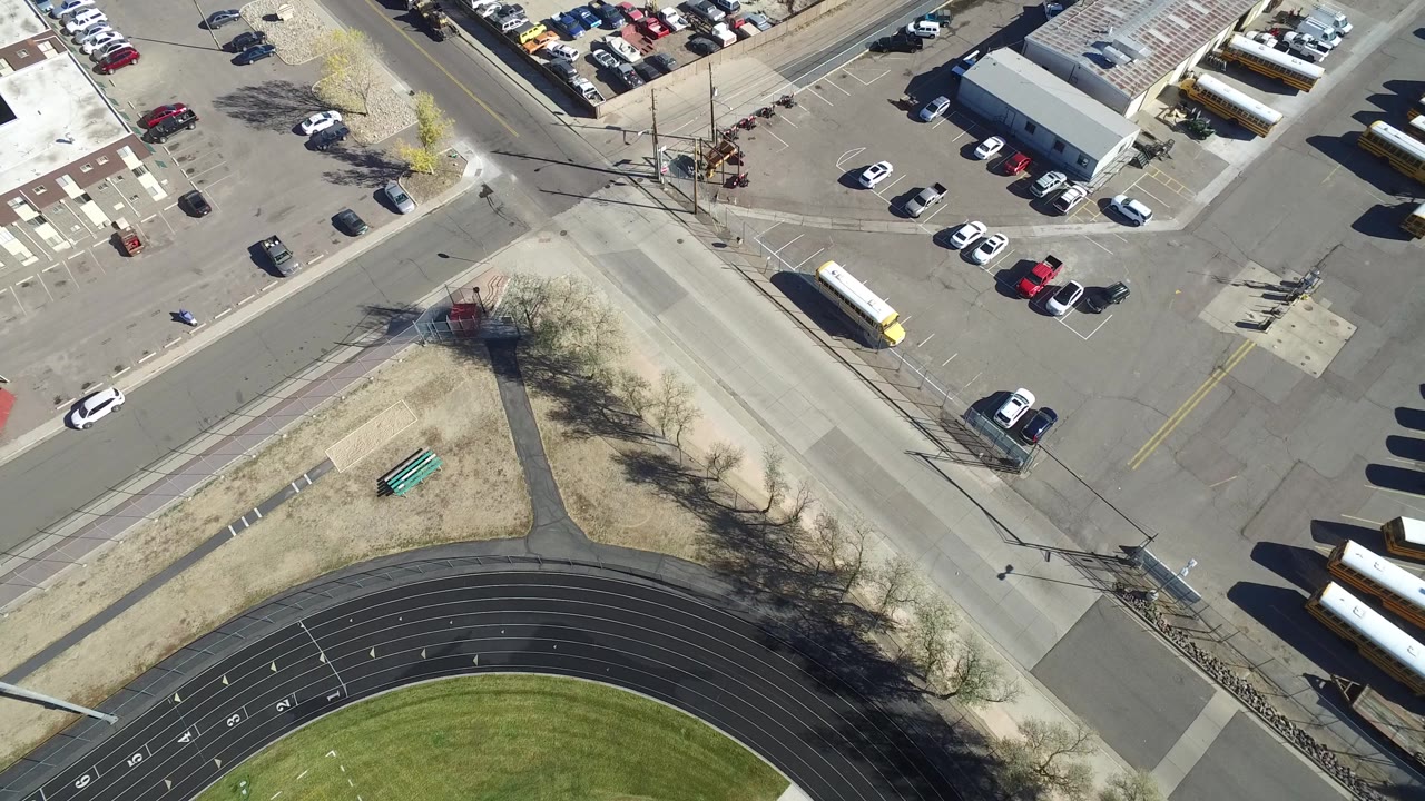 Aerial frame from Hidden Lake, Colorado with buses along the lower right, parked cars near a low building, a road crossing, dry grass, tree shadows, and no visible people.