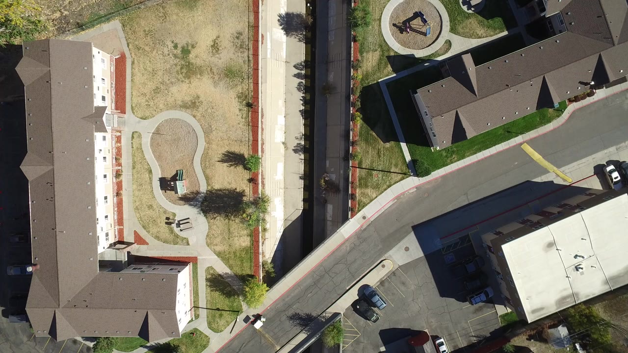 Top-down aerial view of apartment buildings, a curved road, sidewalks, dry grass, and parked cars.