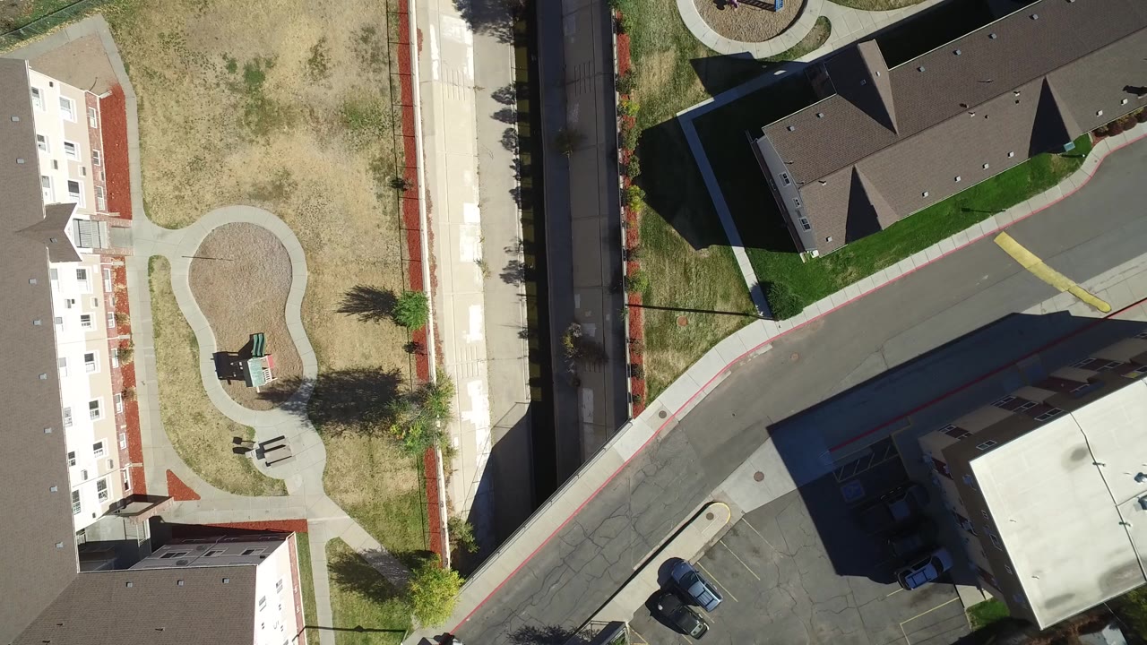 Overhead frame of Hidden Lake, Colorado showing a curved street beside light-colored residential buildings, a dry lawn area, a small looped walkway, trees, and no visible people.