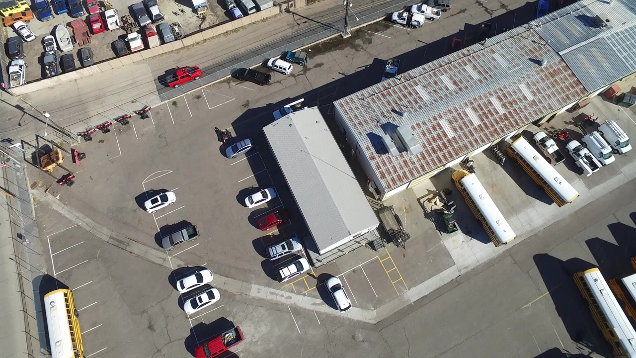 Opening overhead view in Hidden Lake, Colorado showing a commercial lot, parked cars, school buses, a long corrugated roof, and no visible people.