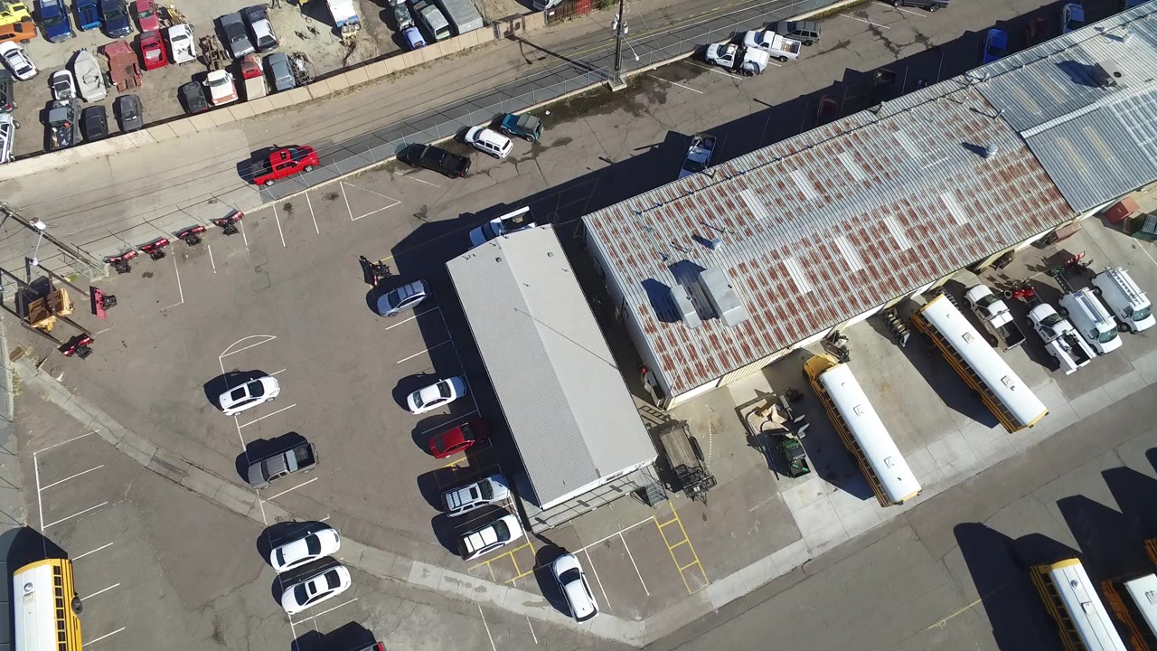 Top-down lot view with metal-roof building, buses, parked cars, and street traffic area.
