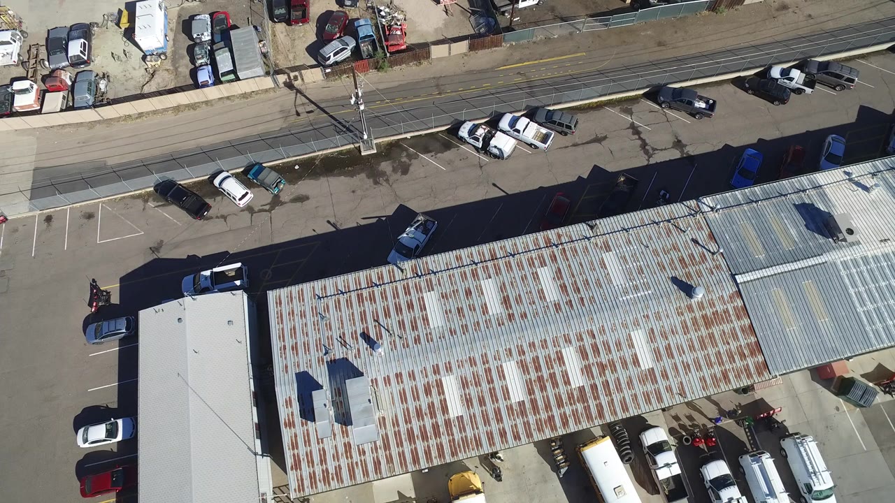 High overhead frame in Hidden Lake, Colorado showing a corrugated commercial roof, parked cars, a nearby road, utility poles, and no visible people.