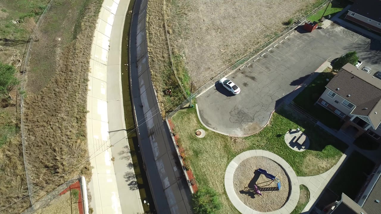 Overhead view of sidewalk, drainage channel, open field, driveway, white car, and house edge.