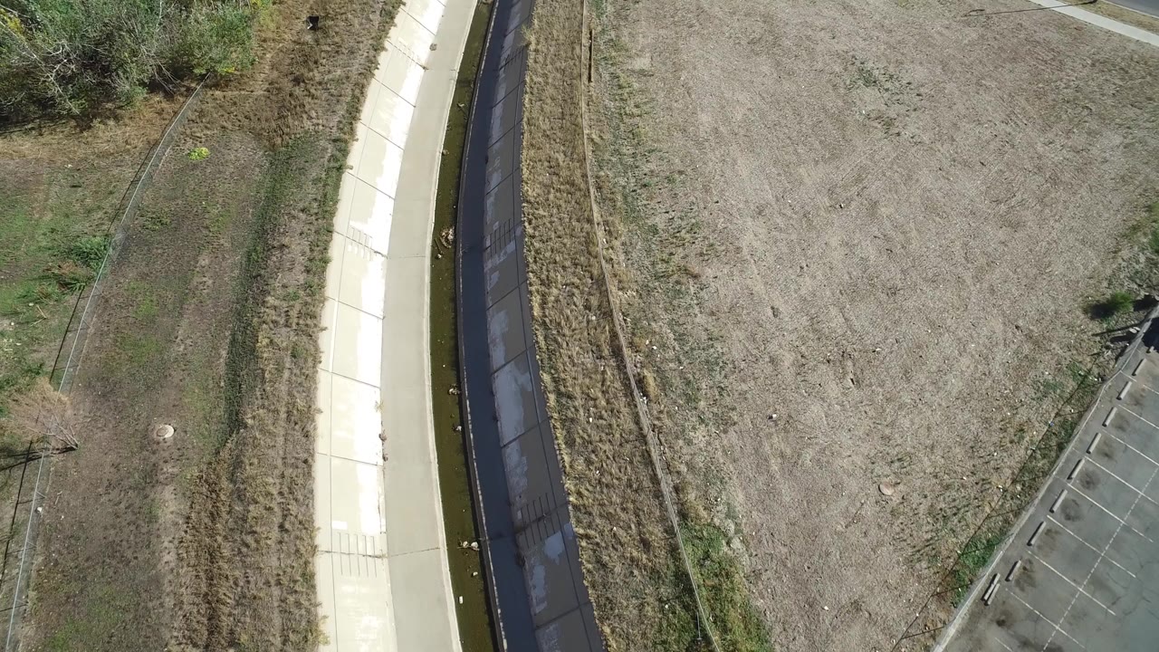 Overhead aerial frame near Hidden Lake, Colorado showing a narrow concrete-lined channel with dark water, dry tan ground, shrubs, fencing, and no visible people.
