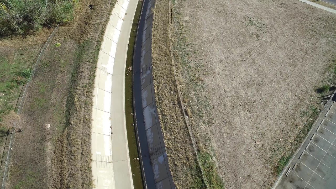 Aerial frame near Hidden Lake, Colorado with the concrete channel along one side, dry open field in the center, a small paved area near the lower edge, and no visible people.