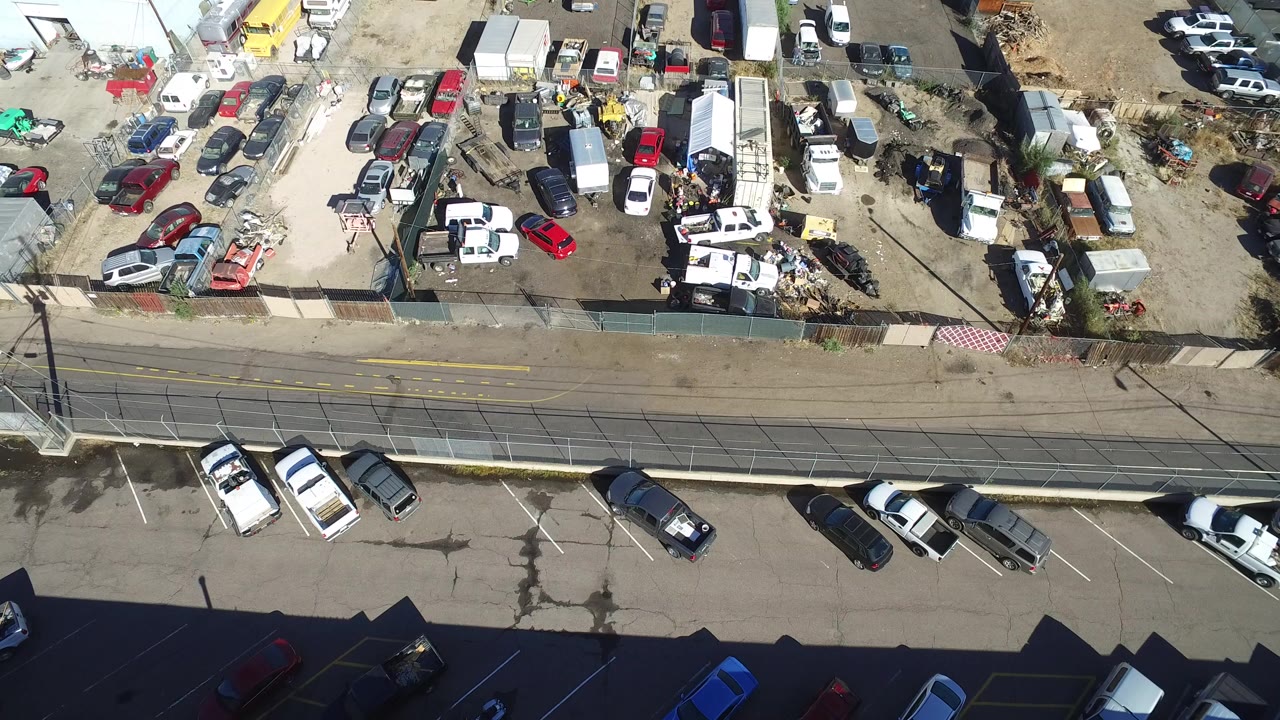 Overhead aerial frame in Hidden Lake, Colorado showing a fenced dirt lot packed with cars, trucks, trailers, and scrap beside a paved road, with no visible people.