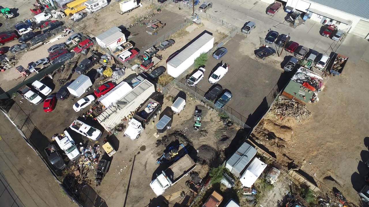 Overhead Hidden Lake, Colorado view showing more of the central dirt parking area, white trailers, parked cars, sheds, debris piles, and no visible people.