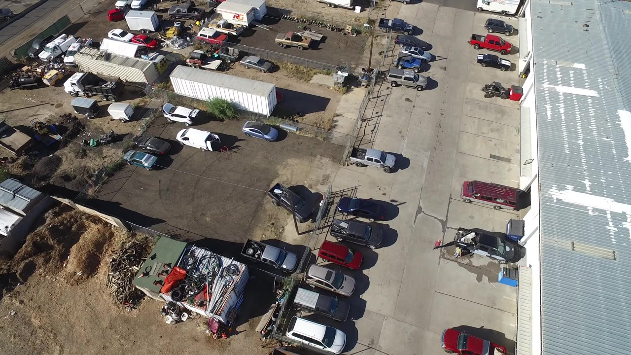 High aerial frame over an industrial lot in Hidden Lake, Colorado, with parked cars, trailers, scrap piles, dirt patches, paved lanes, a pale metal roof, and no visible people.