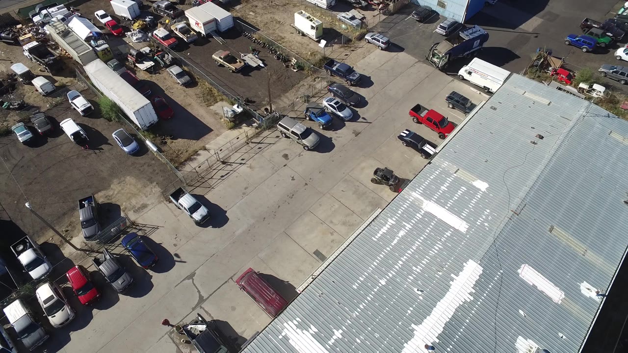 Aerial frame over Hidden Lake, Colorado, with the metal roof taking more of the lower frame, parked cars and trucks along the lot edges, and no visible people.