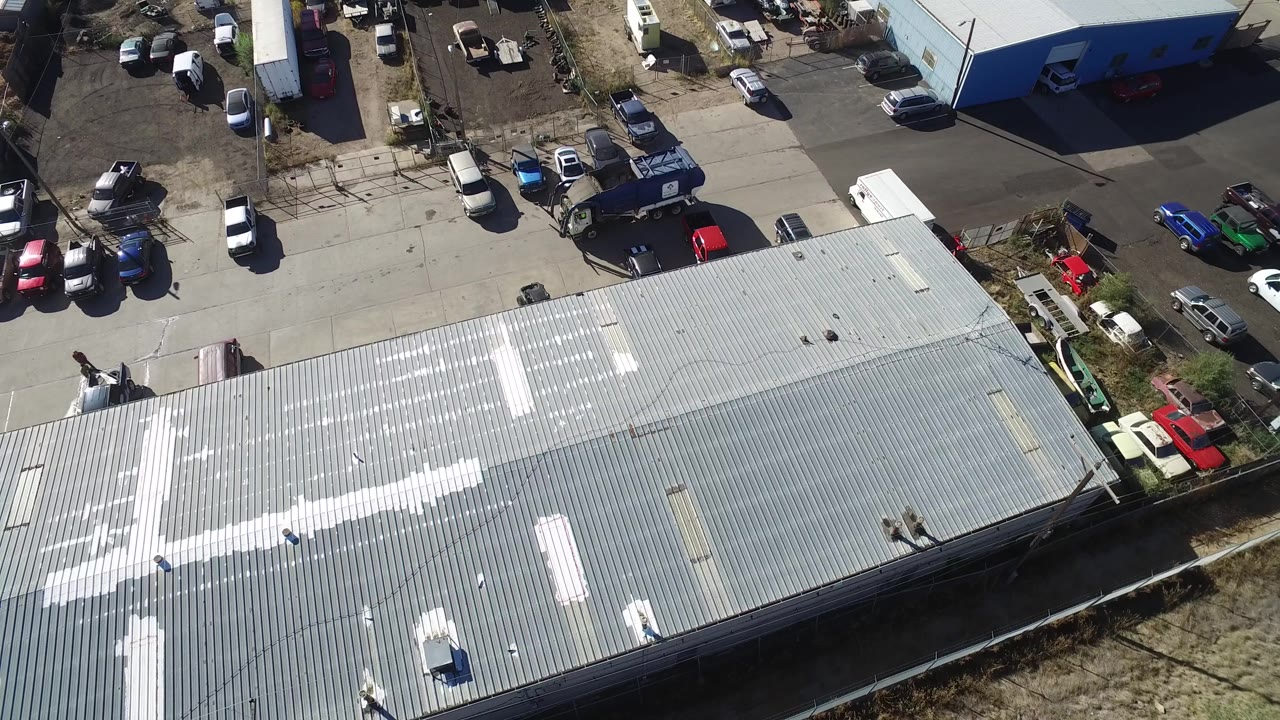 High aerial frame in Hidden Lake, Colorado showing a large ribbed metal roof in the foreground, parking areas with cars beyond it, blue industrial buildings, and no visible people.